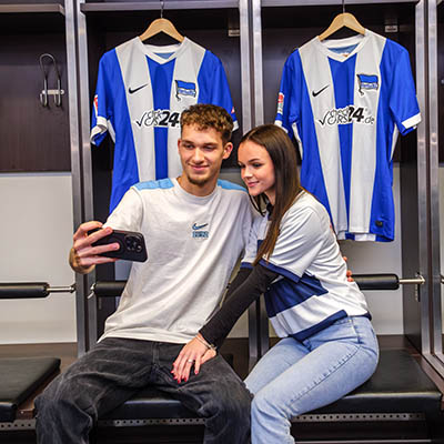 Zwei Personen machen ein Selfie mit Trikots von Hertha BSC.