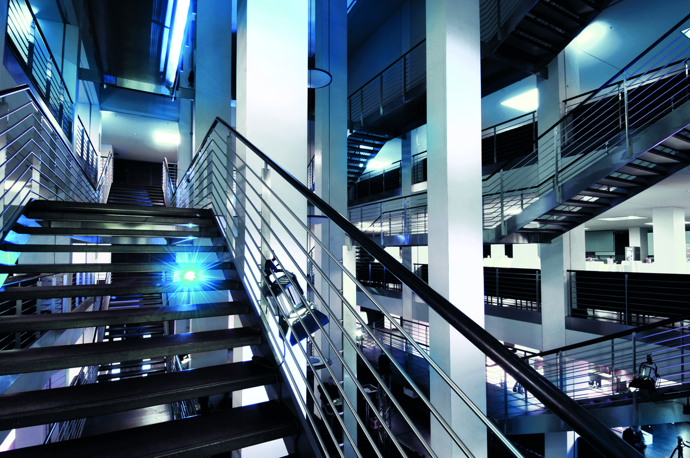 Modernes Atrium im Stadioninnenbereich mit mehreren offenen Treppenfluchten und Geländern aus Edelstahl, dramatisch inszeniert in kaltblauem Licht.