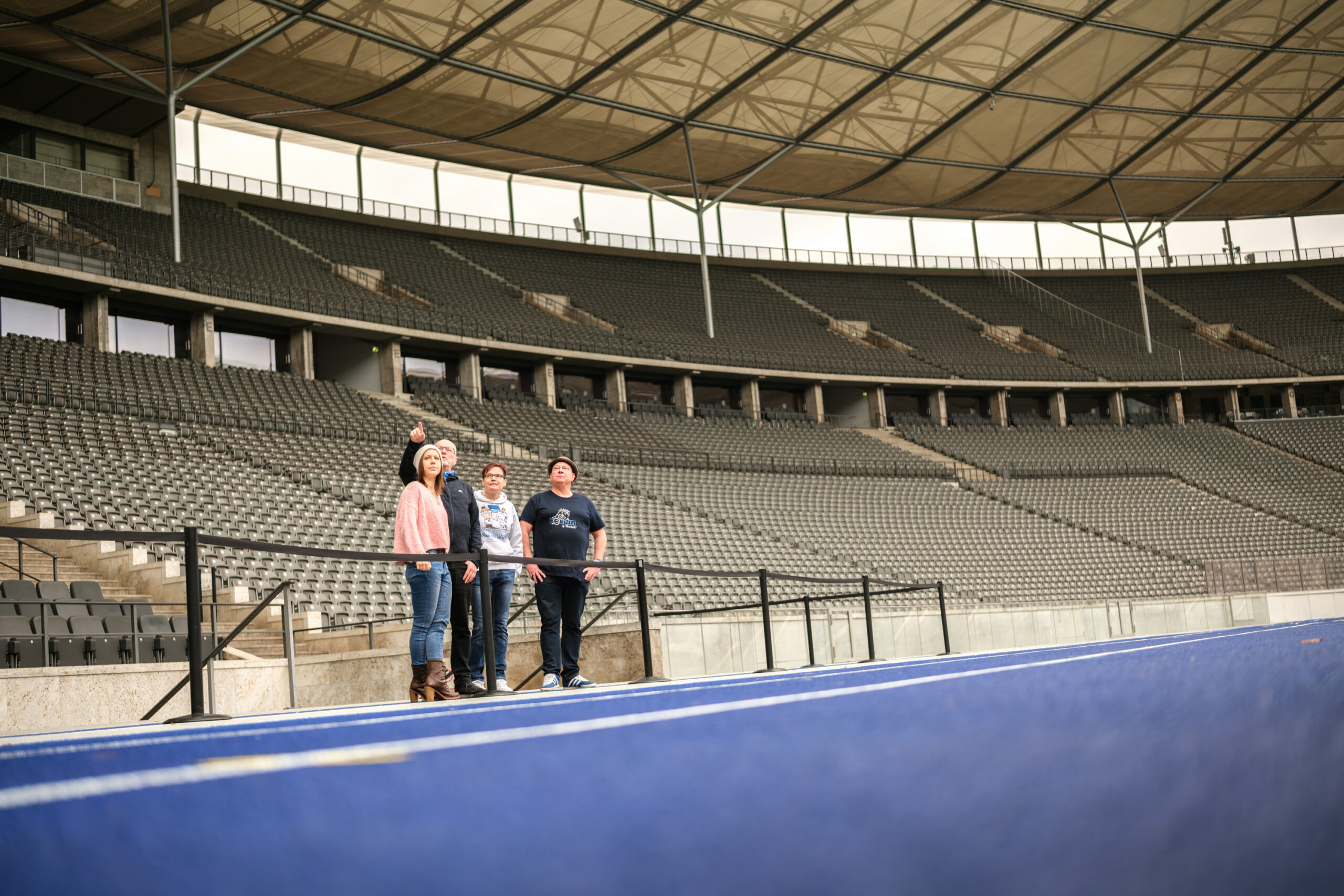Eine kleine Besuchergruppe steht am Rand der blauen Laufbahn im weiten, leeren Olympiastadion. Ein Mann zeigt erklärend hinauf in die großen überdachten Zuschauerränge.