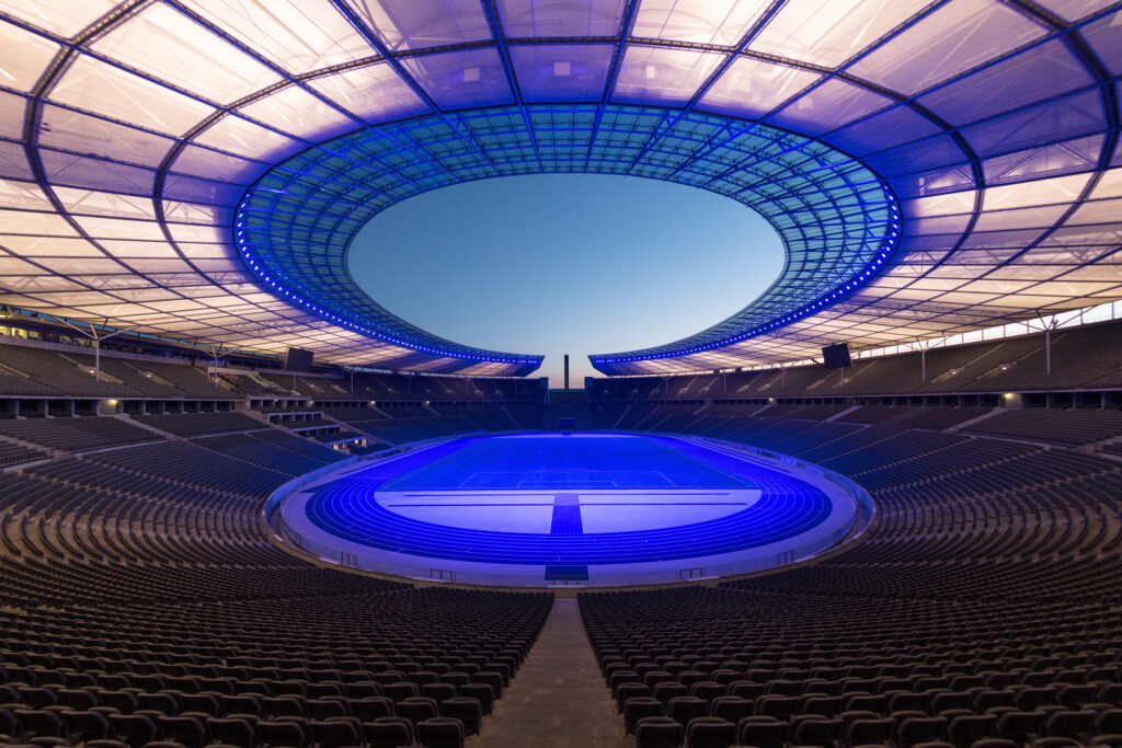Symmetrische Nachtaufnahme des Olympiastadions Berlin mit leuchtend blau inszeniertem Spielfeld, blau illuminierter Dachkonstruktion und dem offenen Marathontor im Hintergrund unter dämmerndem Abendhimmel.