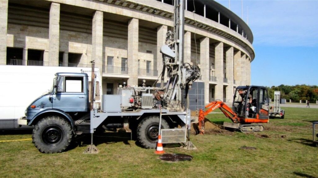 Bohrarbeiten vor der historischen Kulisse des Olympiastadions Berlin mit einer mobilen Brunnen- oder Erdbohranlage auf einem Unimog und einem orangefarbenen Minibagger auf der Rasenfläche.