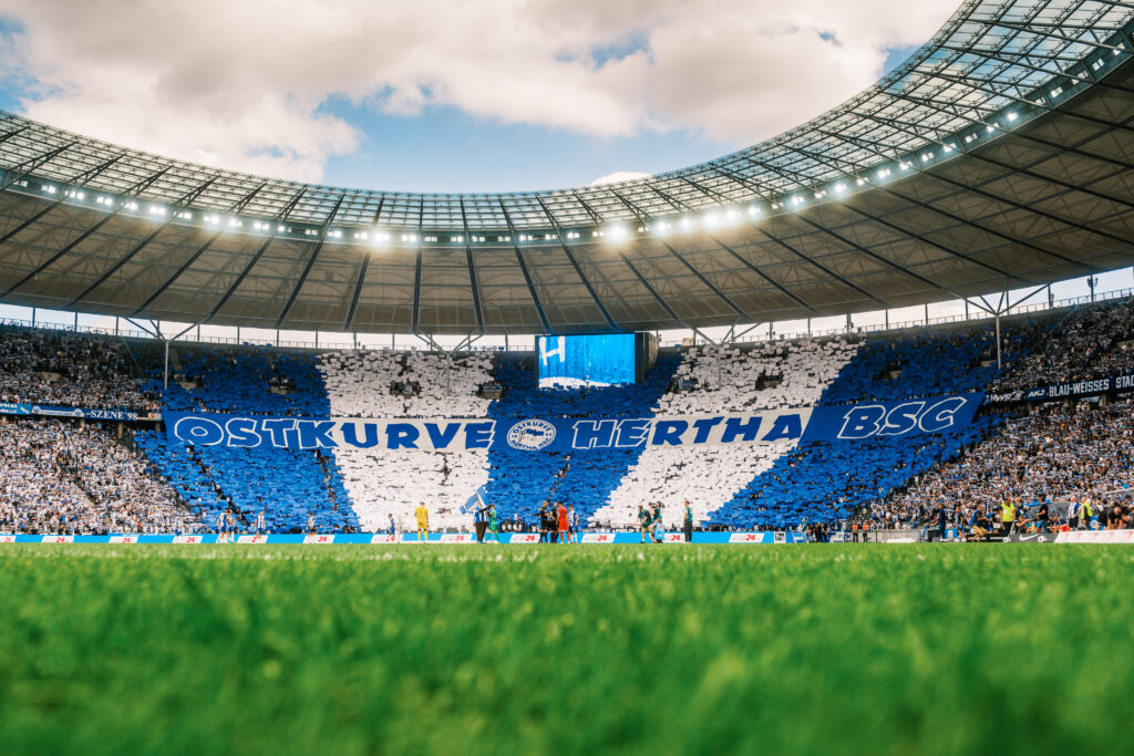 Blick vom Spielfeldrand auf eine große blau-weiße Fanchoreografie in der Ostkurve des Olympiastadions Berlin mit einem riesigen „OSTKURVE HERTHA BSC“-Banner.