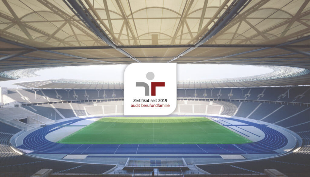 Das Logo des Zertifikats „audit berufundfamilie“ mit dem Zusatz „Zertifikat seit 2019“ liegt als Einblendung über einer Innenansicht des Olympiastadions Berlin mit Rasenfläche und blauer Laufbahn.